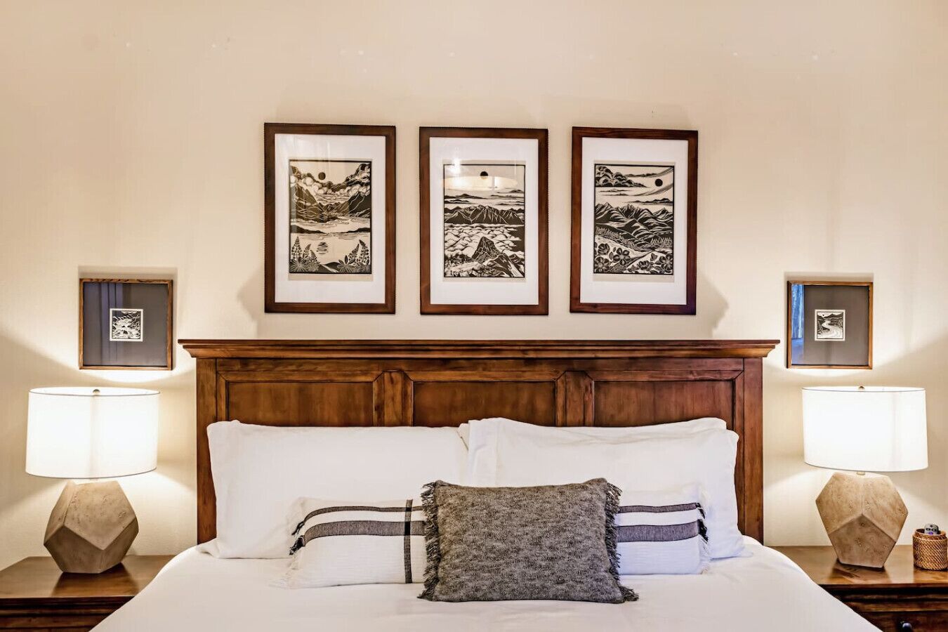 A neatly made bed with white and gray bedding, flanked by two bedside tables with lamps. Above the bed, there are three framed black-and-white landscape paintings on the wall.