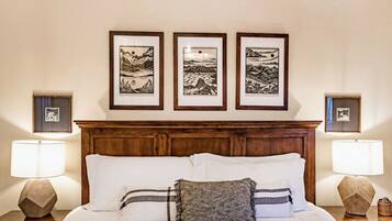 A neatly made bed with white and gray bedding, flanked by two bedside tables with lamps. Above the bed, there are three framed black-and-white landscape paintings on the wall.
