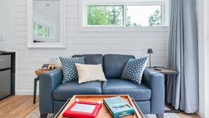 Un salon avec un canapé bleu foncé, deux coussins à motifs, une table basse en bois sur laquelle sont posés des livres, une fenêtre donnant sur des arbres et un rideau gris.