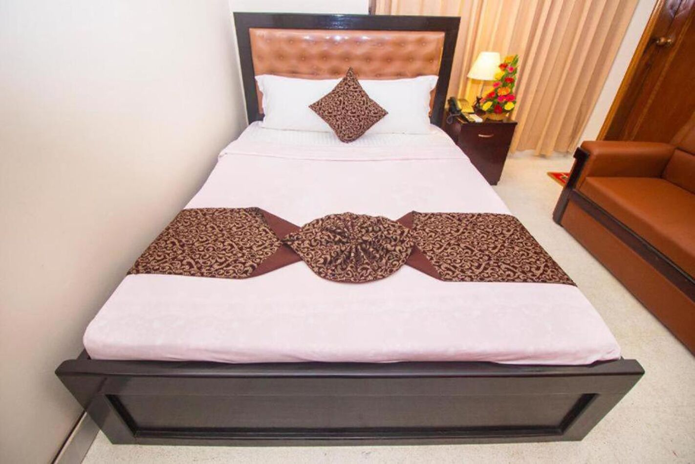 Una cama bien hecha con una colcha estampada, una sola almohada decorativa y una mesita de noche con una lámpara y flores a su lado.