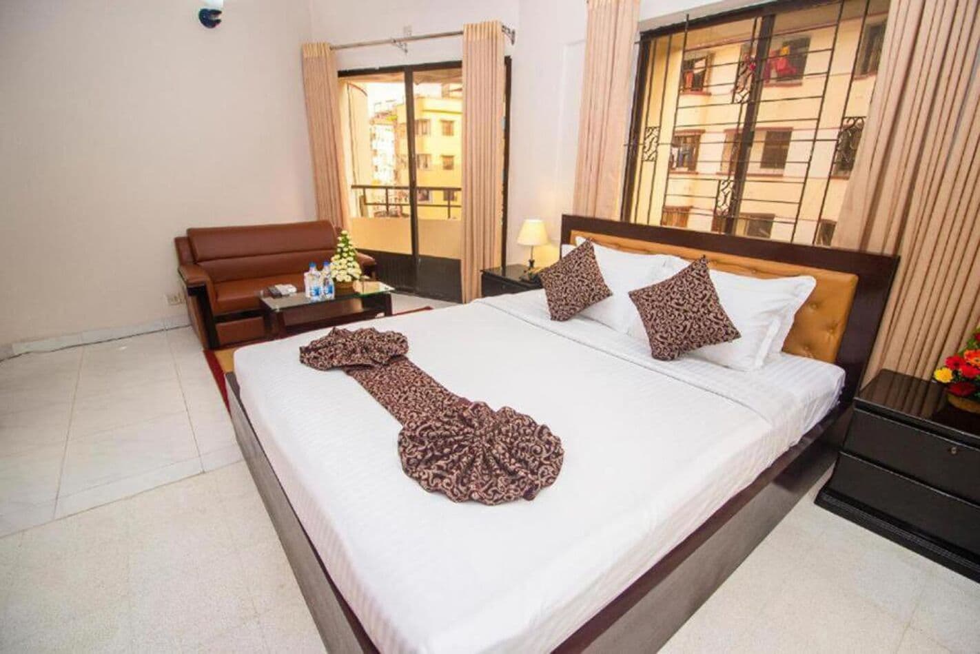 A hotel room with a bed, a nightstand, a lamp, a window with curtains, and a view of buildings outside.