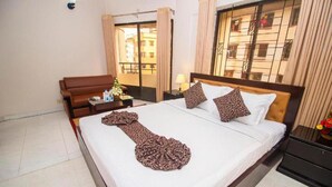 A hotel room with a bed, a nightstand, a lamp, a window with curtains, and a view of buildings outside. - Hotel Sweet Place (Dhaka)