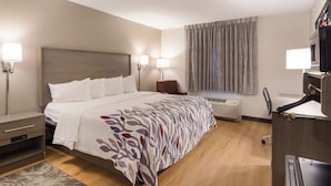 A hotel room with a bed, bedside tables, a desk, a chair, a window with curtains, and an air conditioning unit. - Red Roof Inn Williamsburg North (Williamsburg)
