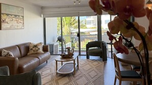 Ein geräumiges Wohnzimmer mit einer braunen Ledersofa, einem runden Holztisch und einem grauen Sessel. Ein Deckenventilator, ein großes Fenster mit Blick ins Freie und ein Gemälde an der Wand sind ebenfalls vorhanden.