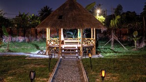 A thatched-roof structure with a wooden deck, surrounded by greenery and a brick wall. - Teko Paradise and Kitchen (Siem Reap)