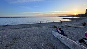 Une plage où l’on voit des gens se promener et s’asseoir sur des troncs d’arbres, un coucher de soleil en arrière-plan et une eau calme.