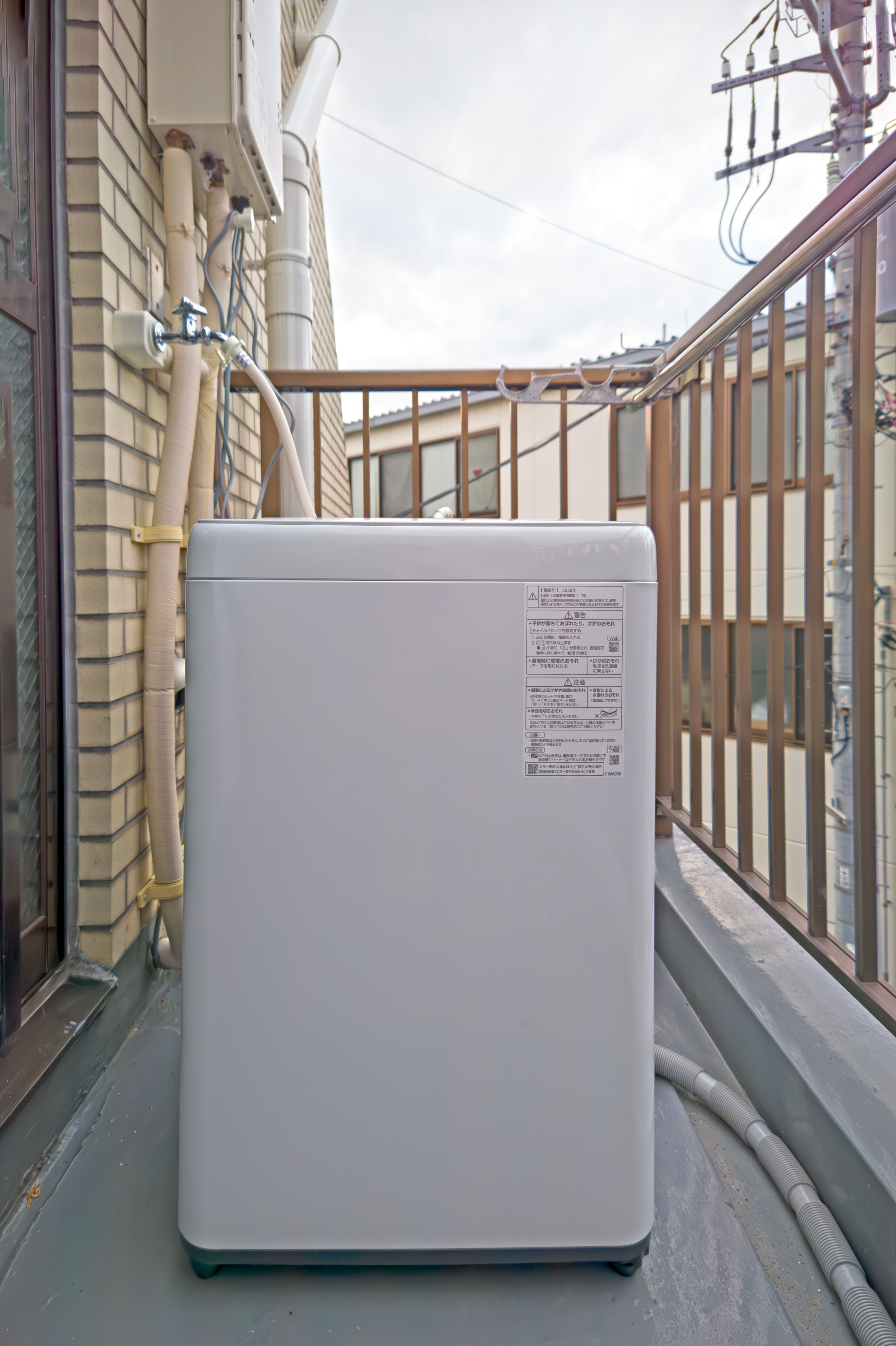 A white portable air conditioner with a label on its side, placed on a balcony with a metal railing and a brick wall in the background.