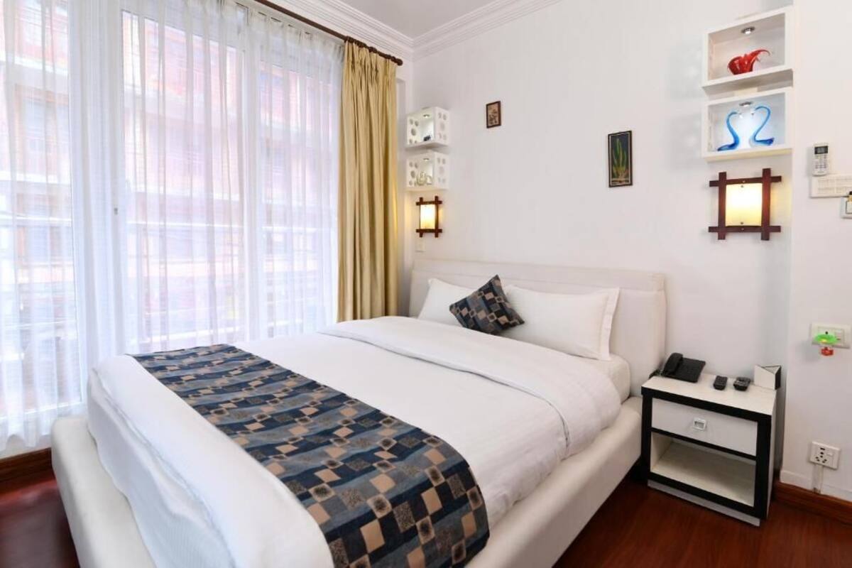 A hotel room with a large bed, a bedside table with a phone and a lamp, and a wall-mounted shelf with decorative items.