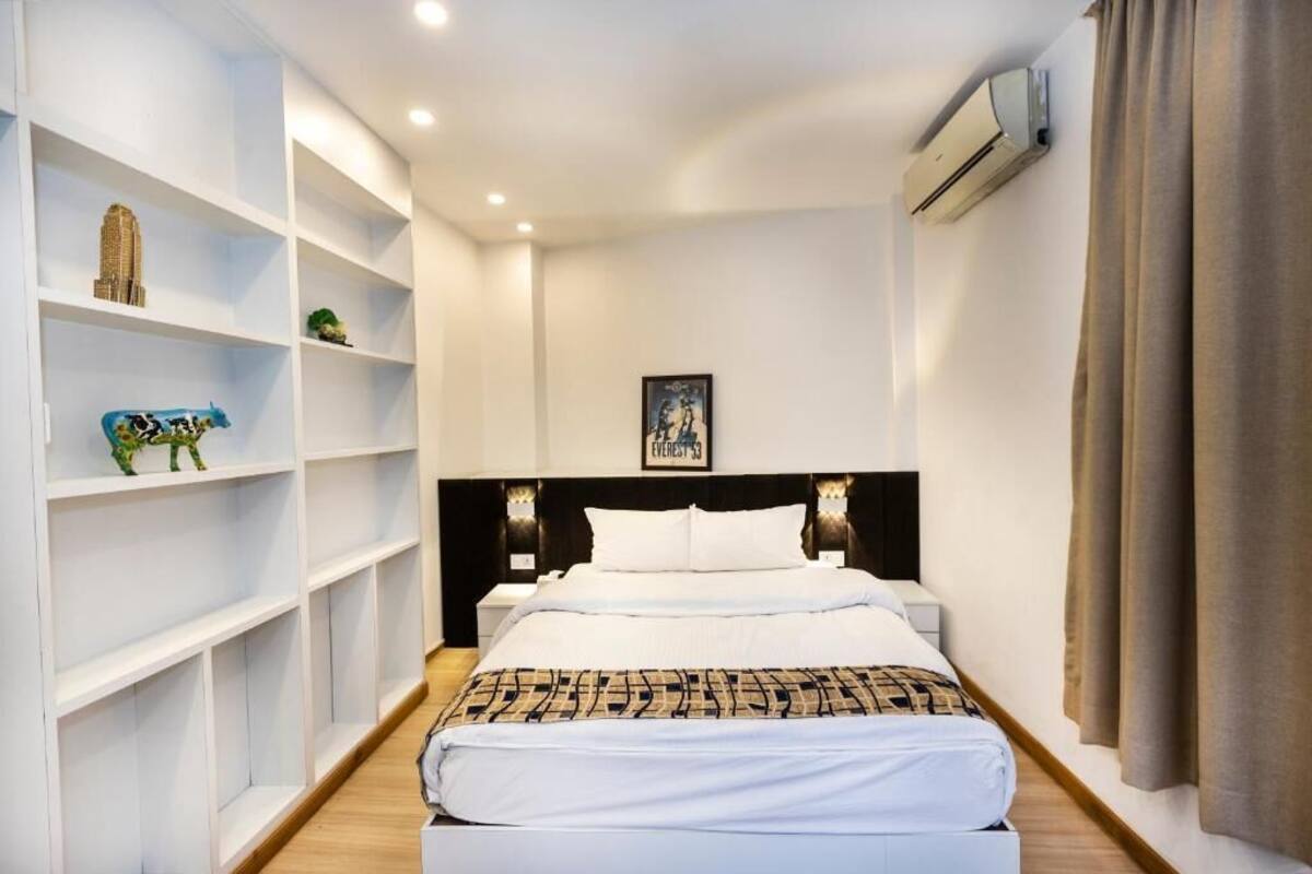 A hotel room with a large bed, white walls, a black headboard, and a shelf with decorative items.