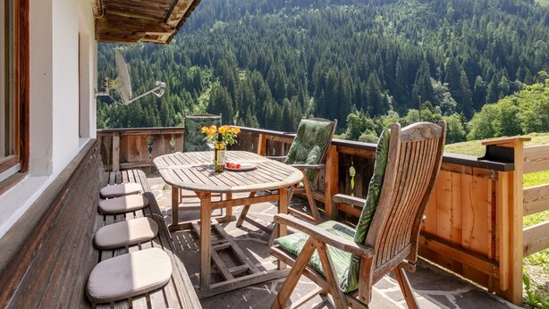 A wooden deck with a table and chairs, a vase with flowers, and a view of a forested mountain. - Farmhouse apartment with mountain view (Alpbach)