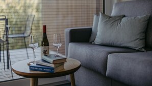 Een moderne woonkamer met een grijze bank, een ronde houten salontafel met boeken en wijnglazen, en een raam met jaloezieën.