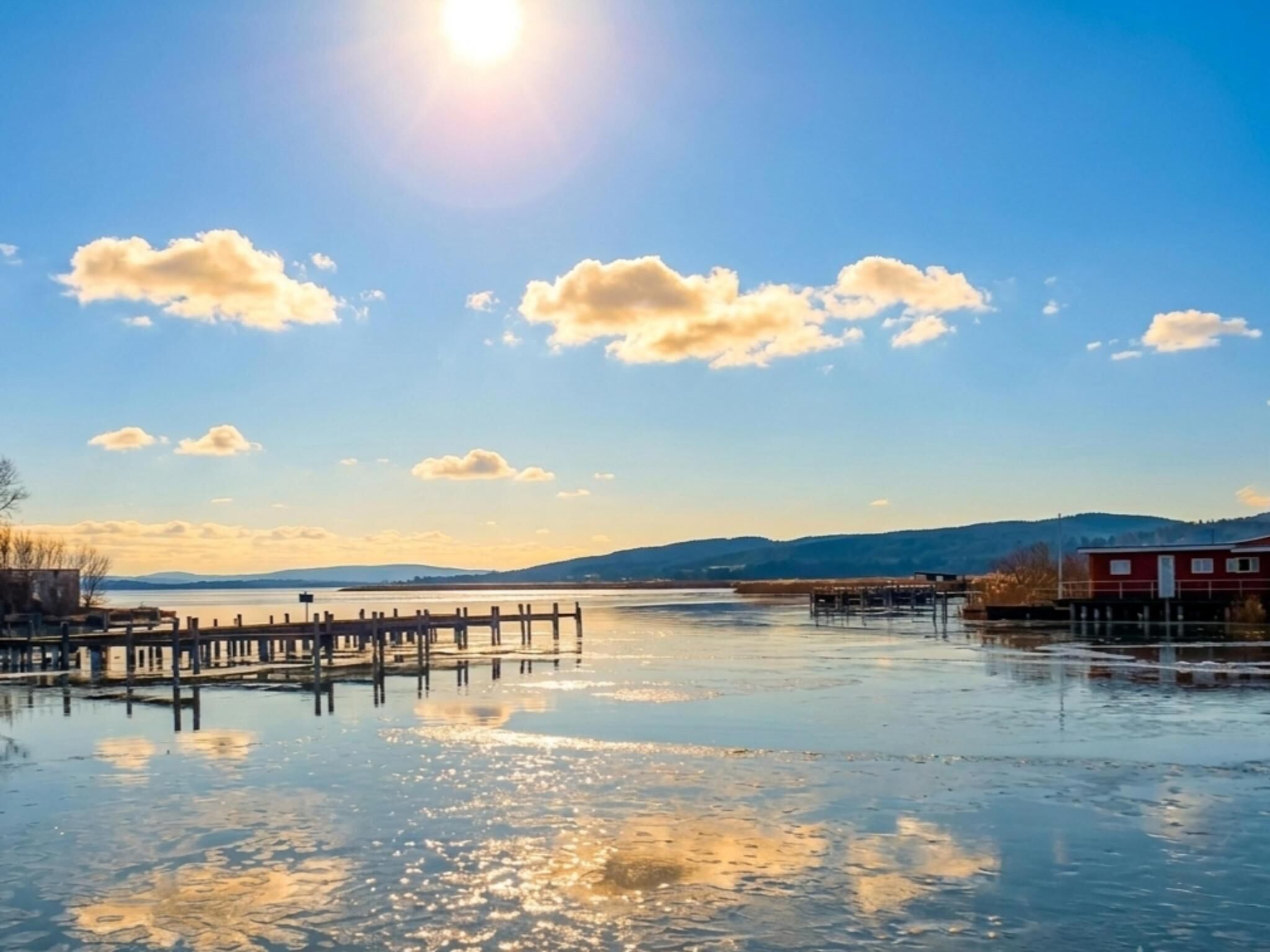 A calm body of water with a wooden dock extending into it, surrounded by a clear sky and distant hills.