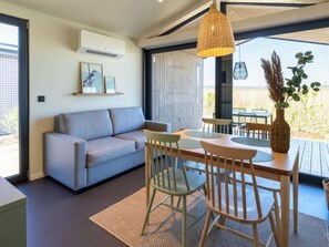 Una sala de estar moderna con un sofá gris, una mesa de comedor de madera y sillas, una lámpara colgante y un ventanal con vistas.