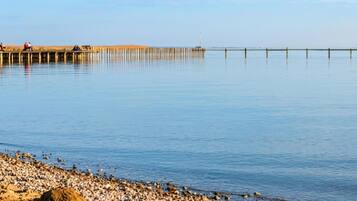 A calm waterfront with a wooden pier extending into the water, a pebbly beach, and a clear sky.