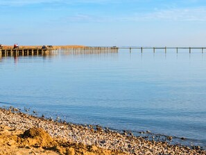 A calm waterfront with a wooden pier extending into the water, a pebbly beach, and a clear sky.