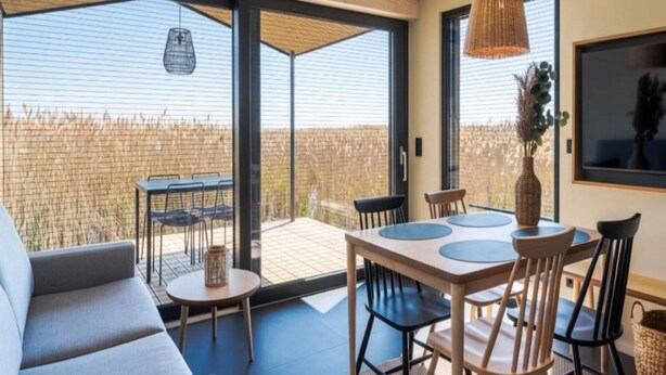 A modern interior with a dining area, a sofa, and a large window overlooking a fenced outdoor area. - Wellness guesthouse by a lake with sauna (Breitenbrunn)