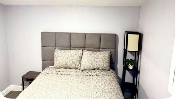 Een slaapkamer met een grijs gestoffeerd hoofdbord, een bed met een patroonbed overtrek, een nachtkastje en een bijzettafeltje met een lamp.