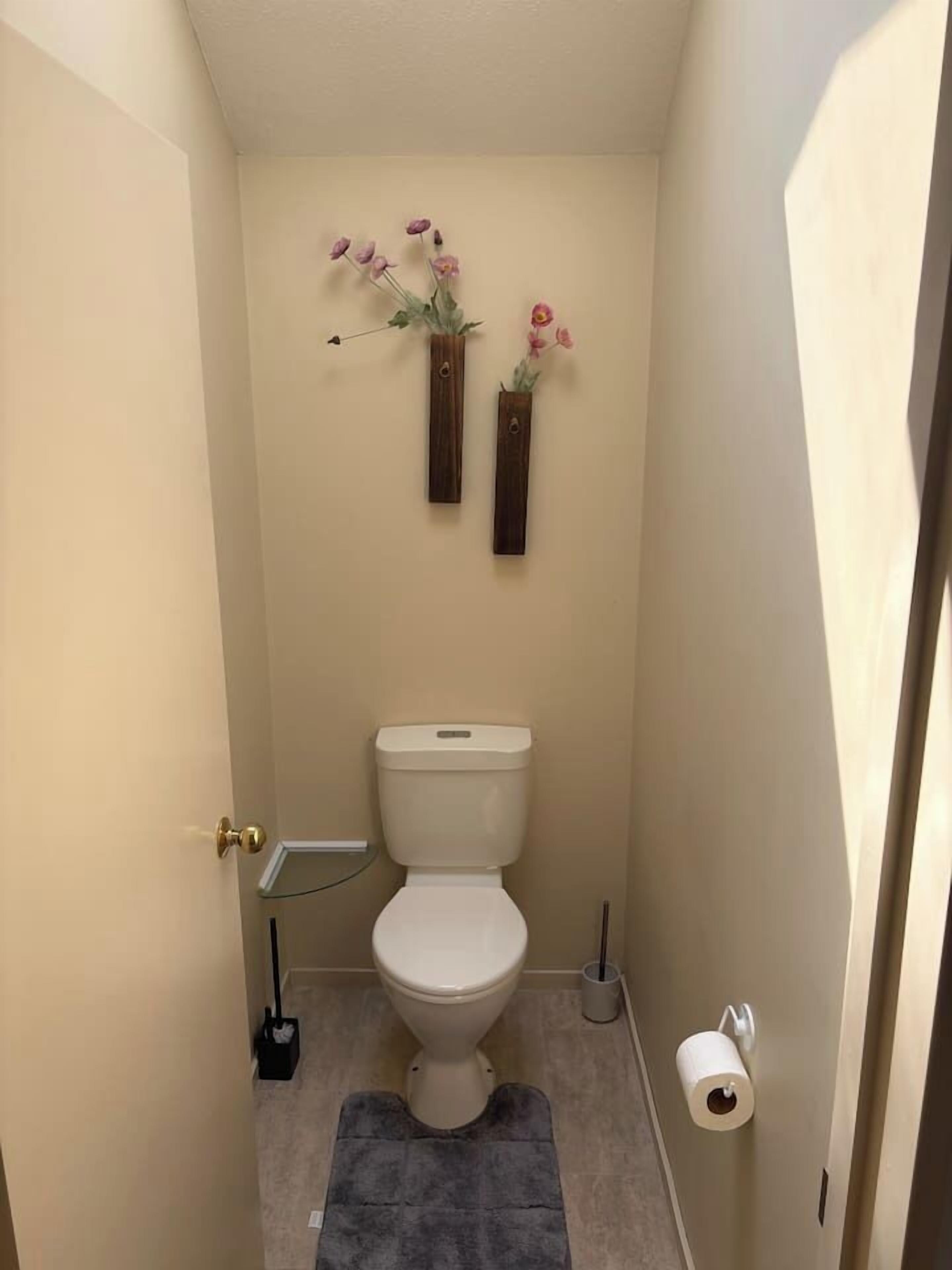 Een compacte badkamer met een toilet, een prullenbak, een toiletrolhouder en twee bloevasen bevestigd aan de muur.