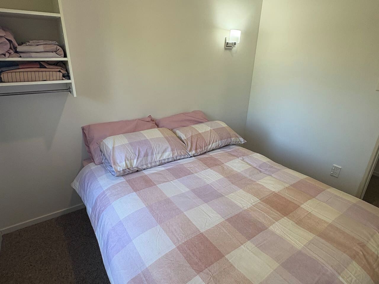 Quarto com cama, prateleira fixada na parede com roupas dobradas e uma tomada.