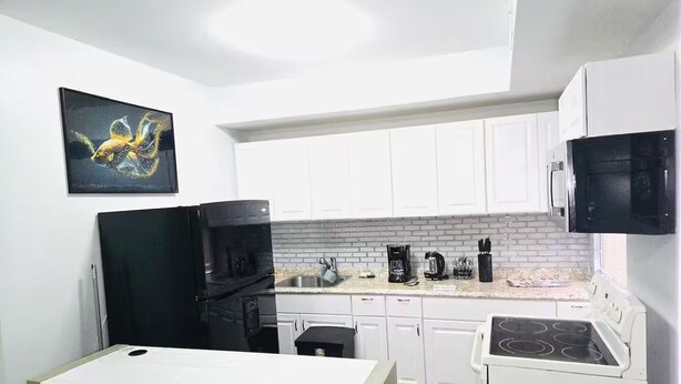 Una cocina compacta con armarios blancos, un refrigerador negro, un microondas, una estufa y una cafetera.