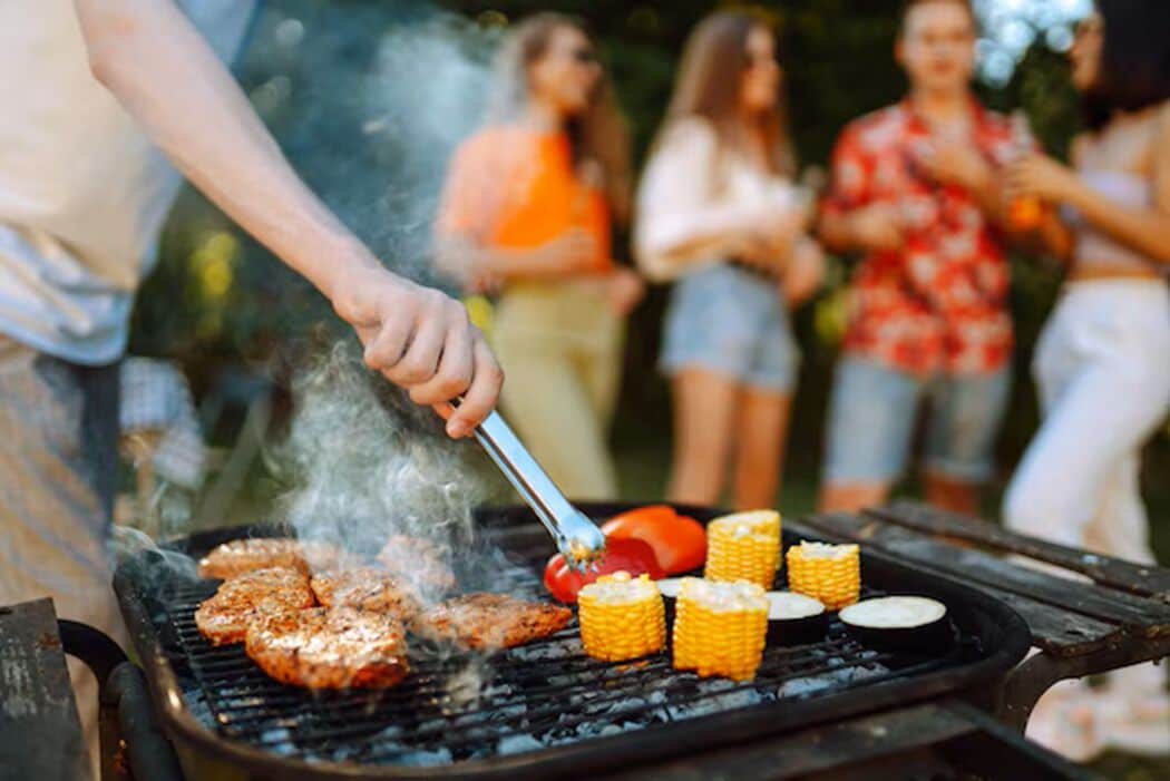 A person grilling food on an outdoor grill with corn on the cob and a red bell pepper.