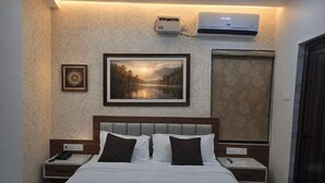 Une chambre d’hôtel avec un lit, deux tables de chevet, une petite table, un tableau encadré, un appareil de climatisation et un rideau à motifs.