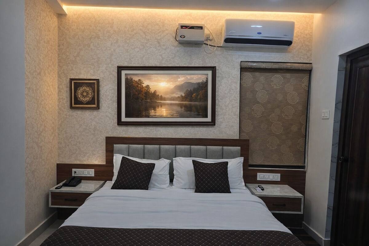 A hotel room with a bed, two bedside tables, a nightstand, a framed picture, an air conditioning unit, and a patterned curtain.