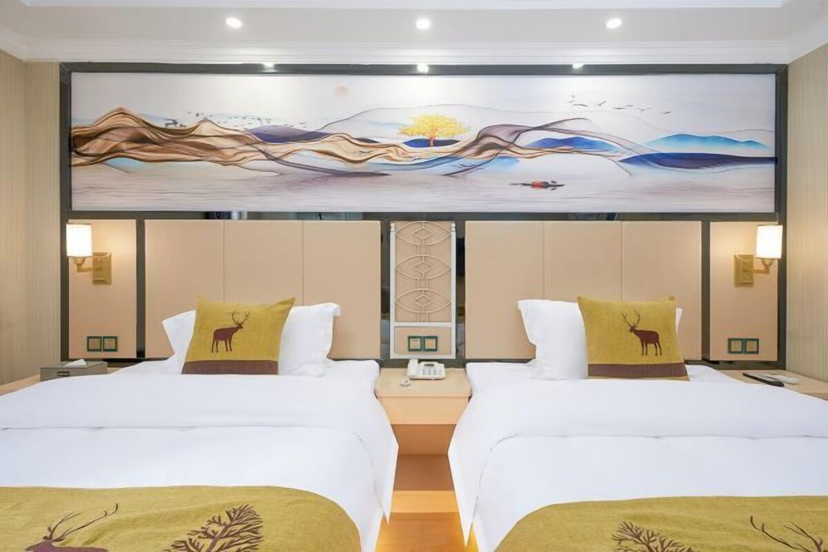 A hotel room with two beds, each with a white bedspread and a pillow featuring a deer design. There is a large painting above the beds depicting a landscape with a tree and mountains.