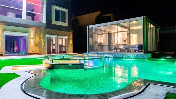 Una piscina con cerramiento de vidrio, un edificio bien iluminado con balcón y una zona ajardinada.