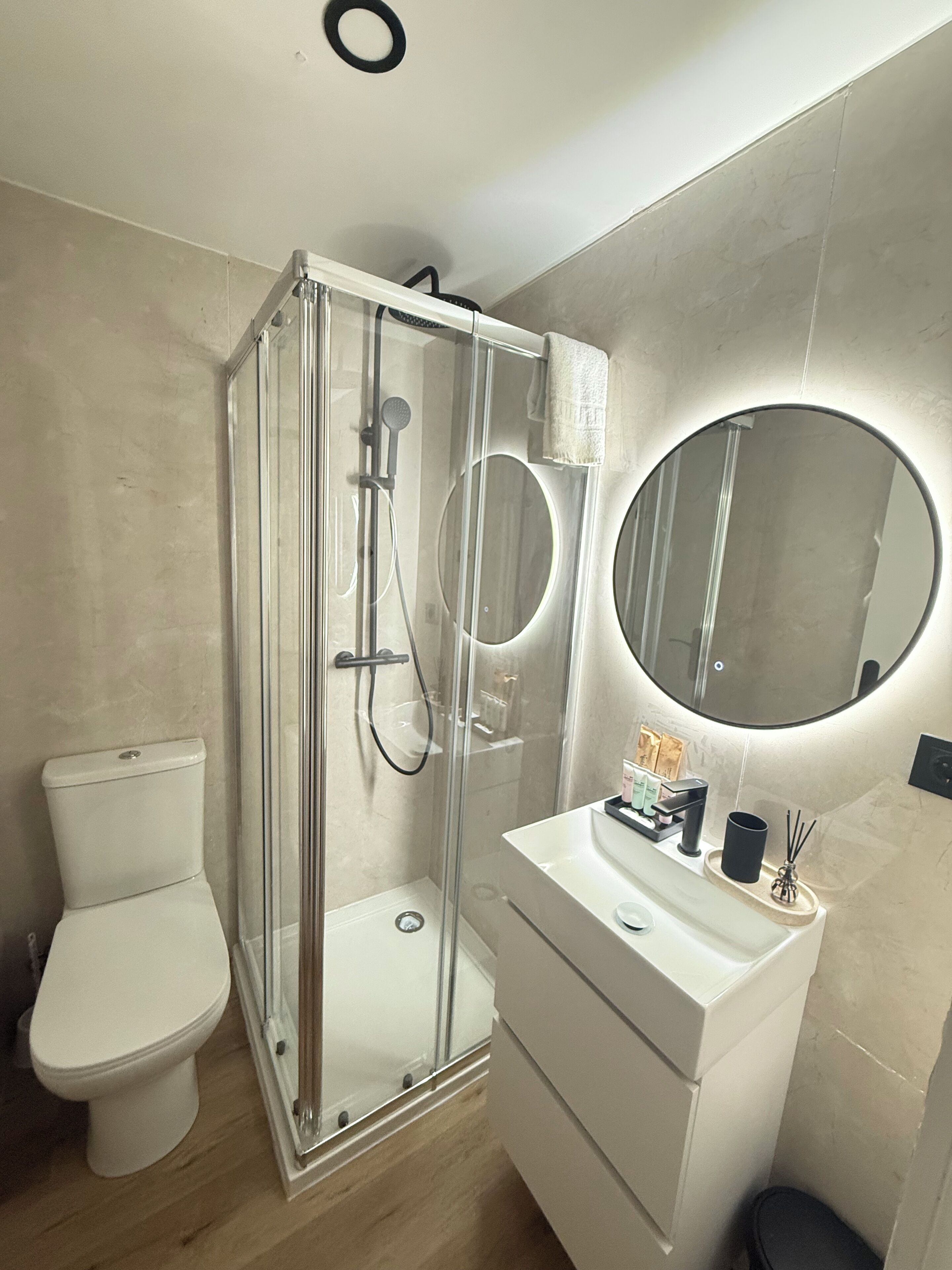 A bathroom with a glass-enclosed shower, a toilet, a sink with a mirror, and a small trash bin.
