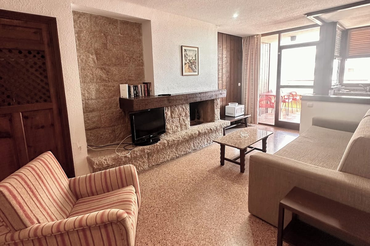 A living room with a stone fireplace, a television, a striped sofa, a coffee table, and a large window.