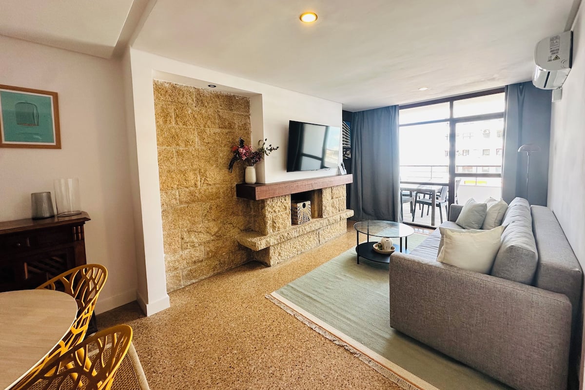 A living room with a stone wall, a flat-screen TV, a sofa, and a dining area with yellow chairs.