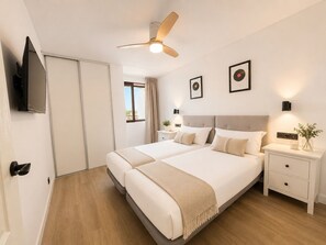 Un lit double avec une tête de lit, des tables de chevet, un ventilateur de plafond, un téléviseur à écran plat et des tableaux encadrés au mur.
