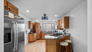 Une cuisine moderne avec des meubles en bois, un réfrigérateur en acier inoxydable, un îlot central doté de tabourets et une fenêtre avec des stores.