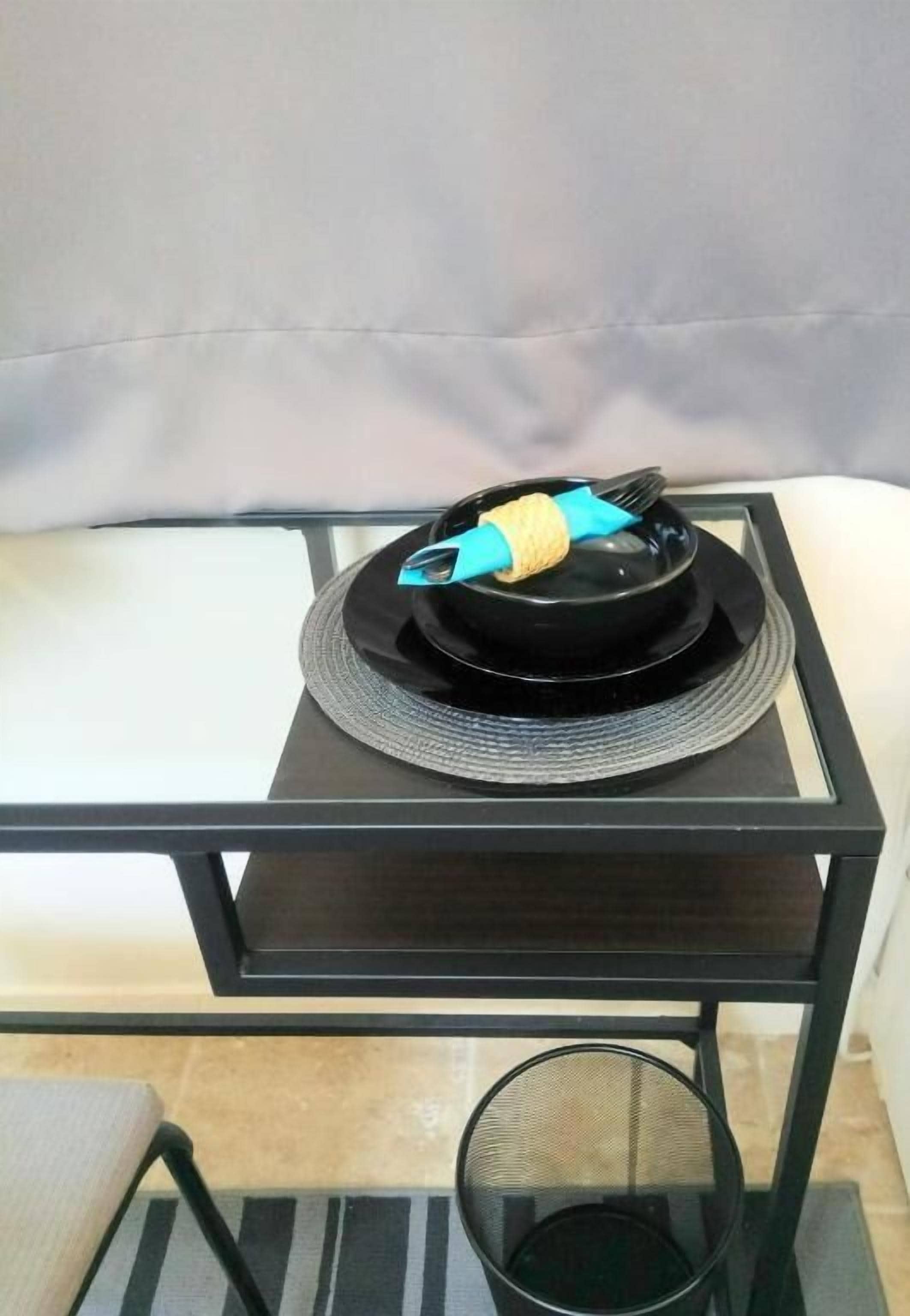 A small black table with a black tray and a bowl on it, placed on a glass table in a room with a grey couch.
