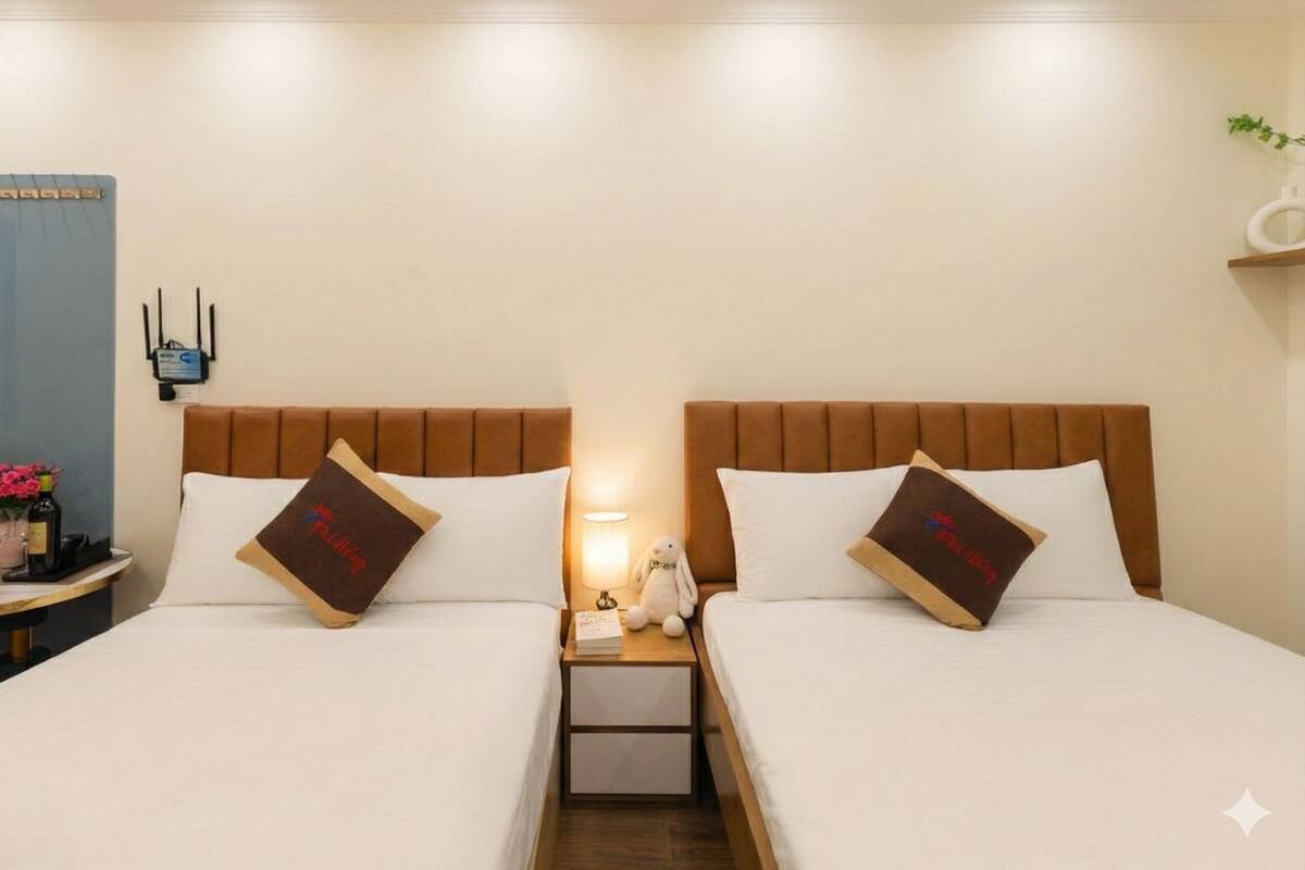 A hotel room with two beds, a nightstand with a lamp, and a small table with a bottle and flowers.