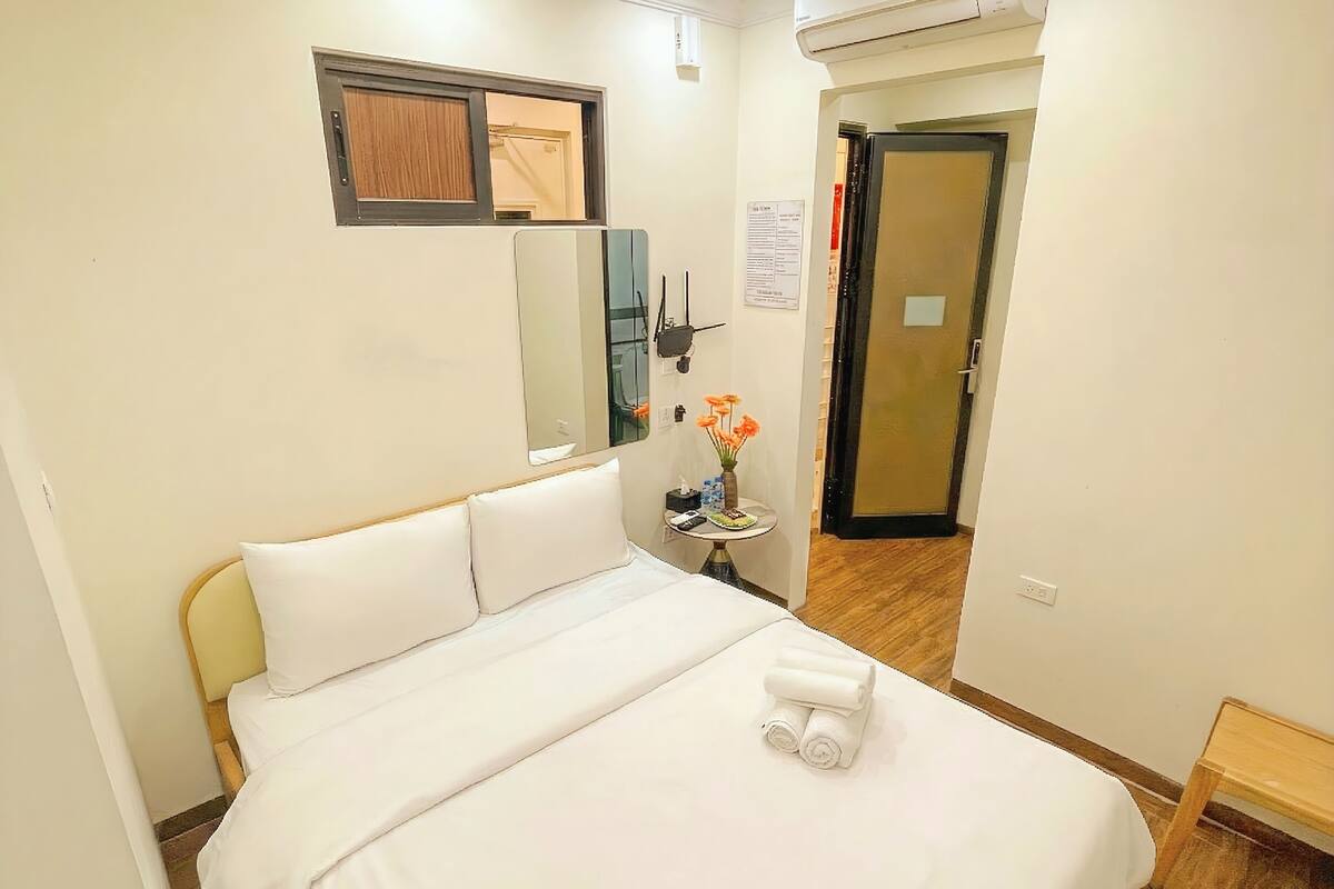 A hotel room with a bed, a small table with a vase of flowers, a mirror, and an air conditioning unit.