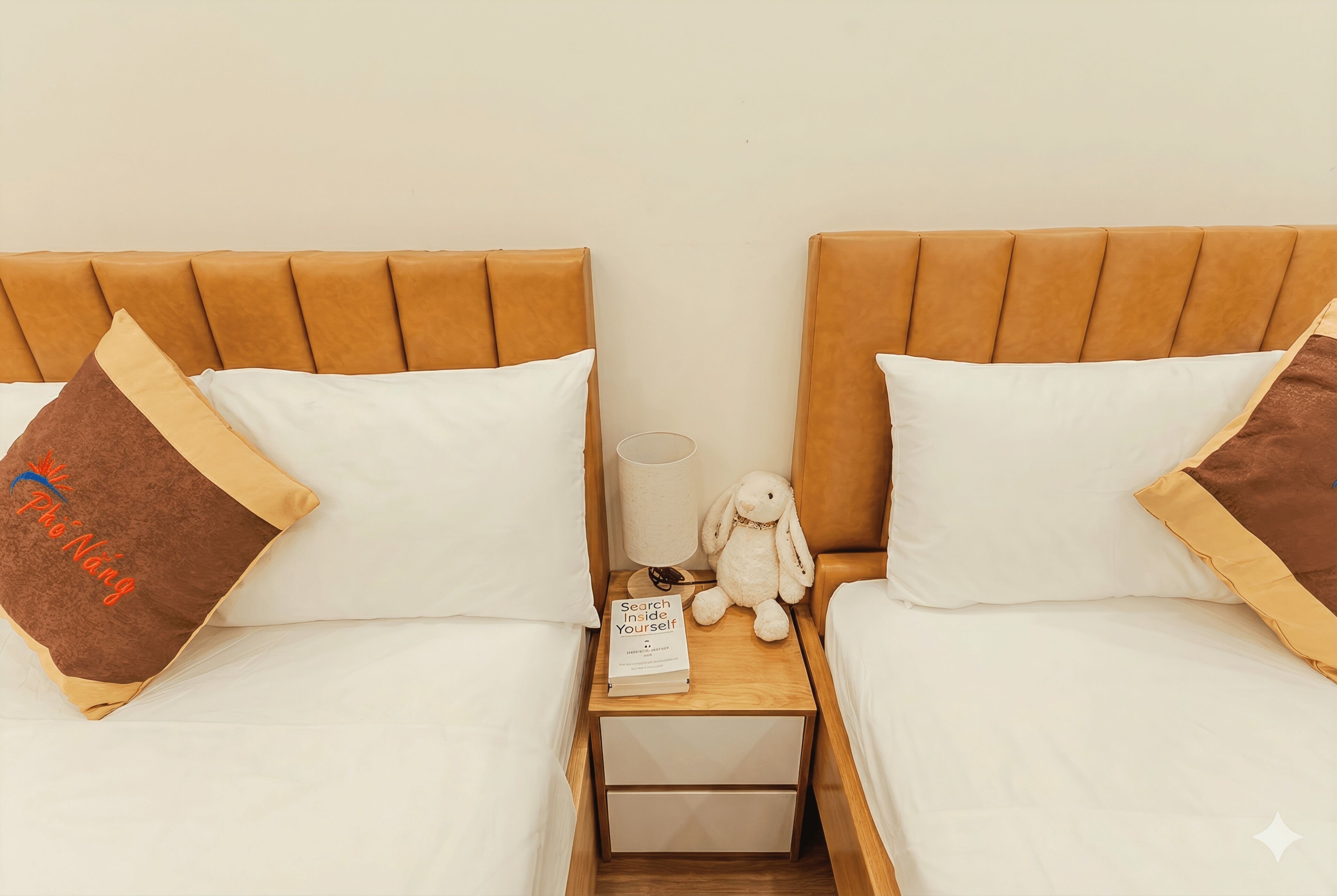 A double bed with a wooden headboard, a small bedside table with a lamp, a stuffed toy, and a book titled "Search Inside Yourself".