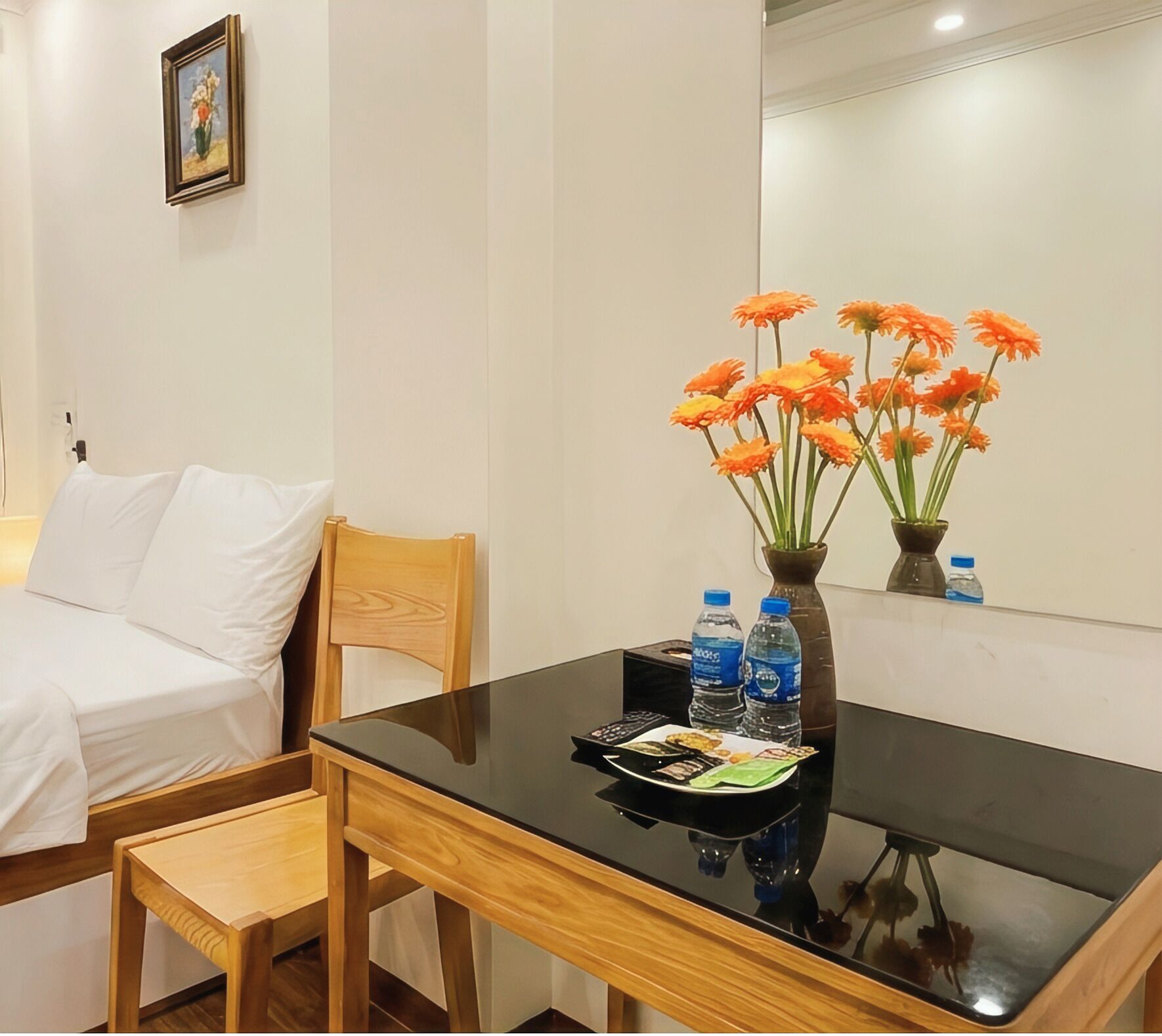 Une chambre d’hôtel avec une chaise en bois, une petite table à la surface noire, un lit blanc et un vase contenant des fleurs oranges.