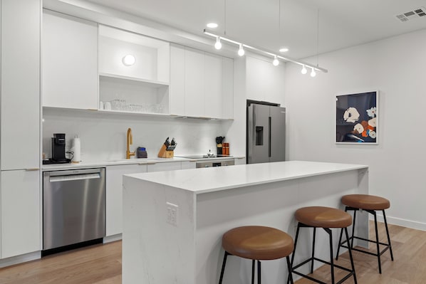 A modern kitchen with white cabinetry, a stainless steel dishwasher, a refrigerator, and a central island with stools. - Experience Zilker & South Lamar from The Code | Private Balcony w/ Pool, Bar (Austin)
