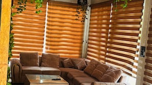 A modern living room with a sectional sofa, a glass coffee table, and wooden blinds. - The Nature Resort (Rishikesh)