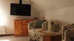 Ein Wohnzimmer mit Flachbildfernseher an der Wand, einem Holzschrank, einem blumigen Sofa und einem kleinen Tisch mit einer Zierpflanze.