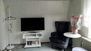 Ein Zimmer mit einem Flachbildfernseher auf einem weißen Schrank, einem schwarzen Sessel, einem kleinen runden Tisch mit Blumen in einer Vase und einem weißen Kleiderschrank.