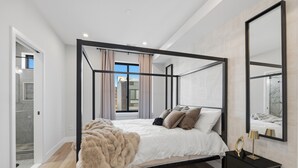 Ein Bett mit Baldachin, ausgestattet mit einer weißen Daunendecke, einer beigen Tagesdecke und mehreren Kissen. Ein schwarzer Nachttisch mit Lampe und Spiegel steht neben dem Bett.