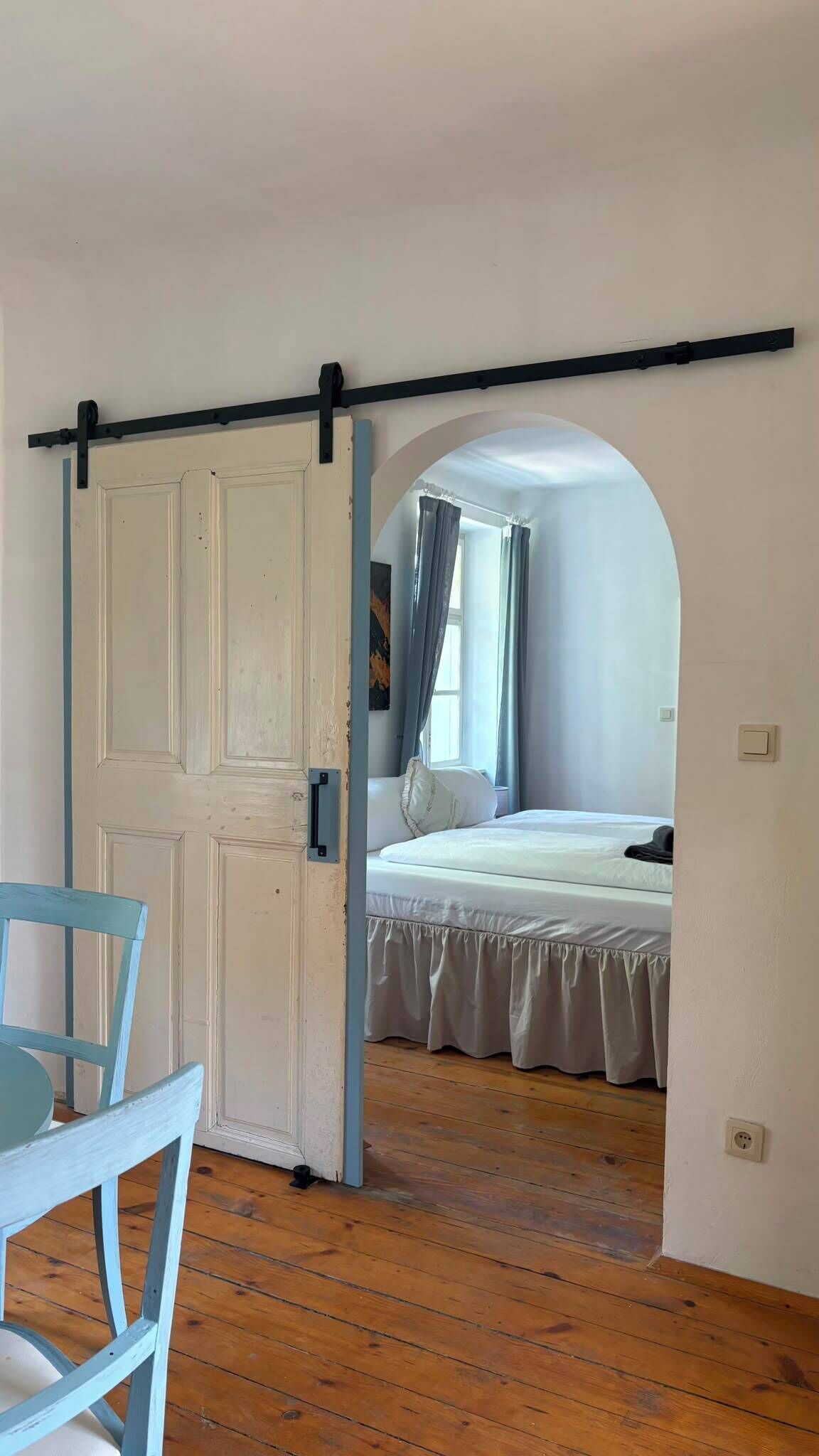 Een kamer met houten vloer, een bed met witte lakens en een stoel.