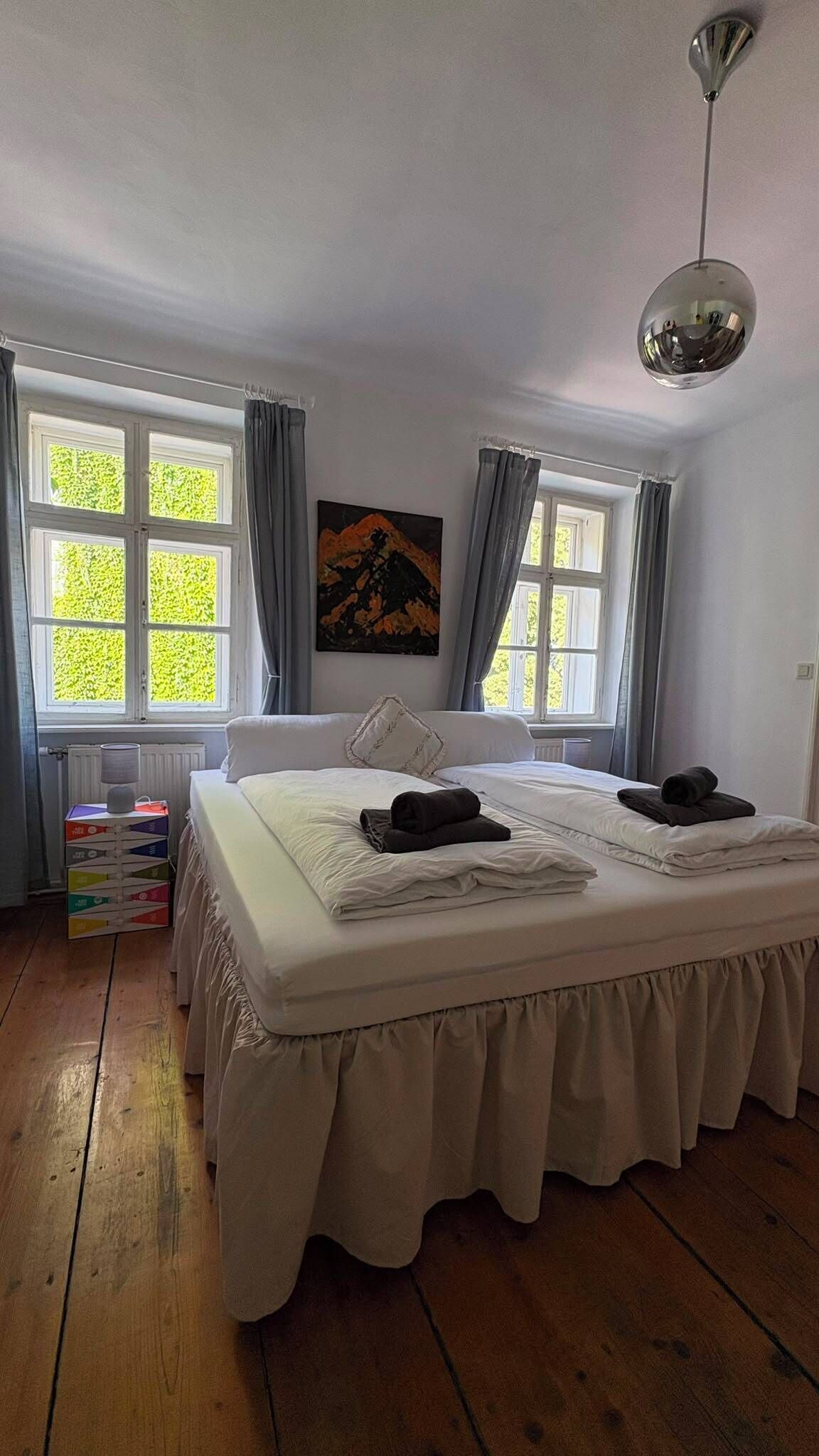 Un dormitorio con una cama grande, dos ventanas con cortinas, un cuadro en la pared y una lámpara en el techo.