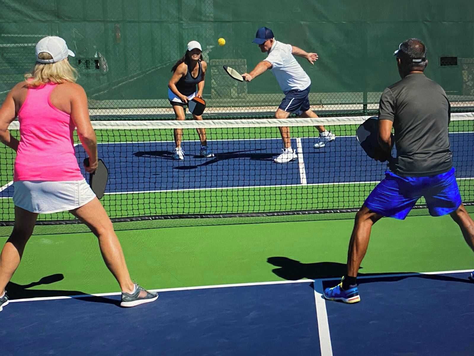 A tennis court with players in motion, a net, and a green fence in the background.