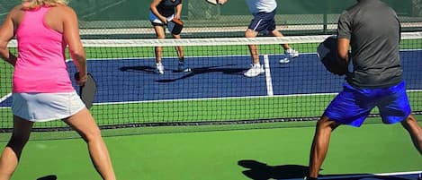 A tennis court with players in motion, a net, and a green fence in the background.