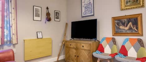 Ein Wohnzimmer mit Fernseher, Holzschrank, zwei Stühle mit bunten Kissen, einer Couch und einem Gemälde an der Wand.