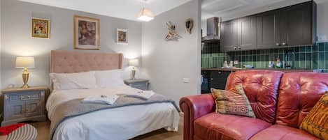 A bedroom with a bed, bedside tables, a red leather sofa, and a kitchen area with cabinets and a green backsplash.